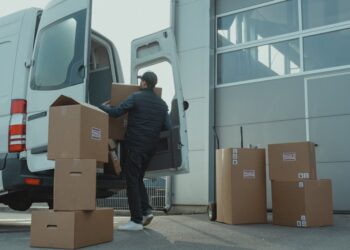 A delivery man unloading cardboard boxes from a van at a warehouse during the day.