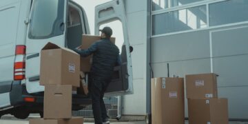A delivery man unloading cardboard boxes from a van at a warehouse during the day.